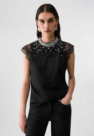 Donna con capelli scuri che indossa grandi orecchini d'argento e una canotta nera senza maniche con maniche in pizzo e perline decorative sullo scollo e sul petto.