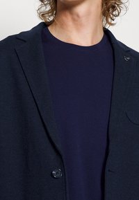 Marinblå blazer med slagkrage, enkel knappstängning och mjukt tyg över en marinblå t-shirt med rund halsringning. Diskret fickdetalj.