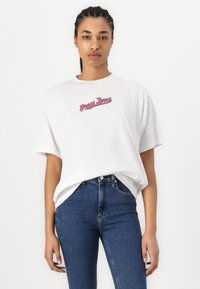 Bijela pamučna majica s okruglim izrezom i kratkim rukavima, koja na prednjoj strani ima izvezeni logo "Guess Jeans" u crvenoj i plavoj boji.