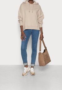Beżowa, oversize'owa bluza z kapturem, niebieskie obcisłe dżinsy, beżowo-białe sneakersy, trzymając torebkę na ramię w kolorze taupe z zewnętrzną kieszenią.