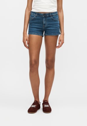 Osoba v modrých rifľových šortkách, bielom nariasenom topu a hnedých semišových teniskách s bielymi šnúrkami stojaca pred jednofarebným pozadím.