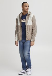 Jachetă cu capușon de culoare bej deschis, cu accente crem, hanorac bleumarin, blugi din denim albastri și adidași albi cu dungi bleumarin. Material neted, ușor.