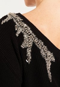 Pull en tricot noir avec un décolleté en V profond, orné de perles argentées décoratives et d'ornements le long des épaules et du col.