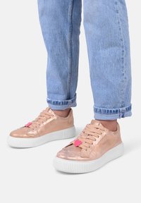 Sneaker rosa oro con finitura metallica testurizzata, suola in gomma bianca e accenti a forma di cuore rosa sui lacci. Indossate con jeans azzurri risvoltati.