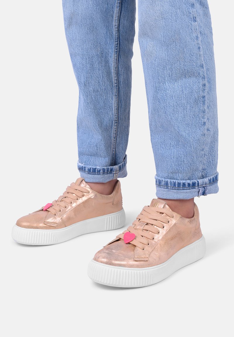 Roségoldene Sneaker mit strukturiertem, metallischem Finish, weißer Gummisohle und rosa herzförmigen Akzenten an den Schnürsenkeln. Getragen mit hochgeschnittenen hellblauen Jeans.
