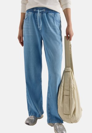 Personne portant un pantalon jogger en denim bleu clair et des baskets grises, tenant à côté d'elle une housse de guitare beige rembourrée.