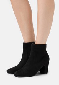 Czarne zamszowe botki za kostkę na słupku, o gładkiej teksturze i eleganckim, minimalistycznym designie. Zamek błyskawiczny z tyłu.