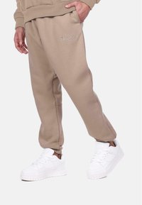 Pantalons de jogging beiges en tissu doux, offrant une coupe confortable et des poches latérales, associés à des baskets blanches volumineuses.