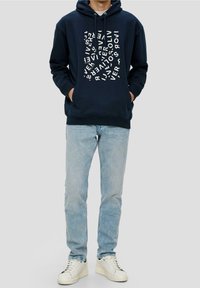 Hombre con sudadera azul marino con texto distorsionado en blanco, jeans azul claro y zapatillas blancas, de pie con las manos en los bolsillos.