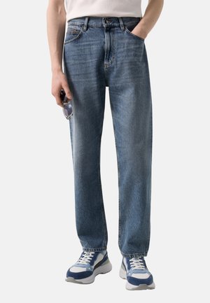 Person iført afslappede blå jeans, hvide og blå sneakers, holder solbriller i venstre hånd, højre hånd i lommen, hvid skjorte delvist synlig.