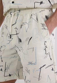Helle beigefarbene Shorts mit einem Zugbandbund, die mit abstrakten schwarzen und blauen Linienmustern versehen sind und den Markennamen "Lagerfeld" auf der Vorderseite tragen.