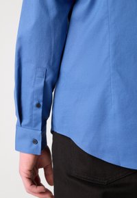 Camicia blu a maniche lunghe con finitura strutturata, dotata di polsini con bottone e orlo curvo. Indossata sopra pantaloni scuri.