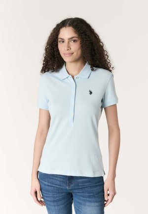 Donna con capelli ricci che indossa una polo a maniche corte azzurro chiaro e jeans blu, in piedi davanti a uno sfondo chiaro e semplice.