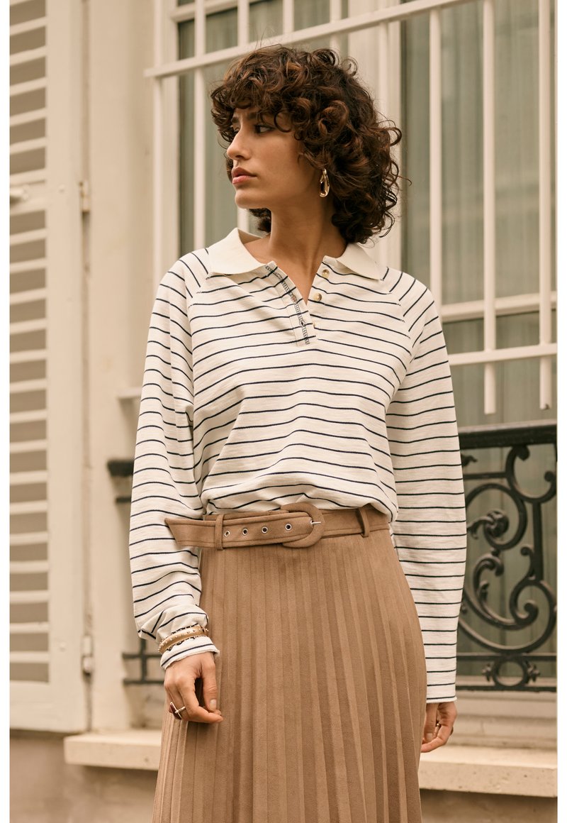 Donna con capelli ricci che indossa orecchini a cerchio dorati, una camicia a righe a maniche lunghe e una gonna plissettata color sabbia con cintura, in piedi all'aperto vicino a una finestra.