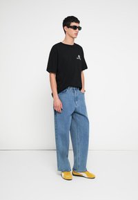 Czarny t-shirt z krótkim rękawem z logo, jasnoniebieskie luźne dżinsy oraz żółte trampki. Dżinsy mają luźny krój i klasyczną teksturę denimu.