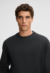 Czarna bluza z okrągłym dekoltem, wykonana z miękkiej, teksturowanej tkaniny. Posiada długie rękawy i luźny krój, bez widocznych logo czy wzorów.