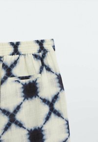 Gros plan sur un pantalon blanc cassé avec un motif tie-dye noir, une ceinture élastique et une unique poche arrière.