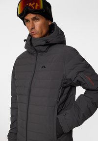 Grå isolerad jacka med huva, med en quiltad design, dragkedja framtill och logga på bröstet och ärmen. Strukturerat tyg och stilren finish.