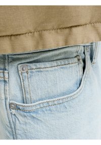 Τσέπη από ανοιχτό μπλε τζιν με μεταλλικά νύχια και πορτοκαλί ραφή κάτω από ένα μπεζ ύφασμα