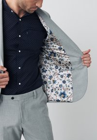 Completo grigio chiaro con interno a fantasia che presenta disegni floreali blu. Camicia blu scuro con sottile motivo a pois e bottoni. Vestibilità su misura.