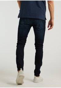 Tmavě modré džíny se štíhlým střihem a jemným vyblednutím, s kapsami na zadní straně a opotřebovanými okraji, doplněné světle barevnými ležérními botami.