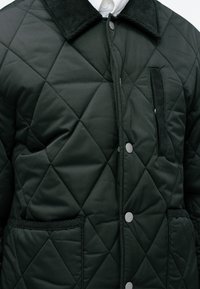 Chaqueta acolchada de un verde oscuro con cuello de pana, botones a presión y un bolsillo con parche. Los acentos texturizados a lo largo de los bordes aportan contraste.