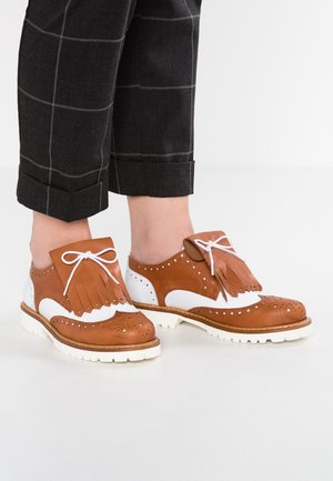 Braune und weiße Brogue-Schuhe mit weißen Schnürsenkeln und Fransen-Details, getragen zu schwarzen karierten Cropped-Hosen.