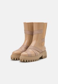Botas de tornozelo à prova d'água em bege com um acabamento brilhante, painéis laterais elásticos e solas de borracha grossas e estriadas. Apresenta a marcação nas correias.