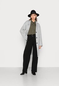 Casaco de lã cinza com um corte descontraído, botões pretos, blusa verde-oliva com um laço, e calças largas pretas combinadas com botas de tornozelo pretas.