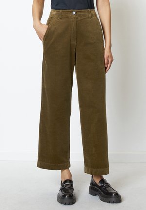 Vrouw draagt olijfgroene wijde ribfluwelen broek, zwarte loafers en een zwarte top, staand tegen een effen witte achtergrond.
