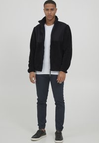 Svart fleece zip-up jakke med en glatt panel, hvit t-skjorte, marineblå bukser med lommer, og svarte joggesko med hvite såler.