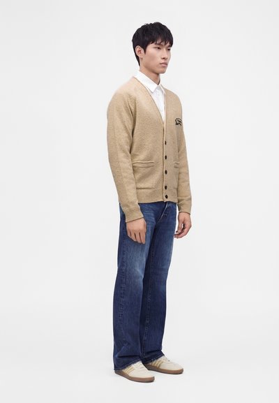 Jeune homme debout portant un cardigan beige boutonné, une chemise blanche, un jean bleu et des baskets blanches sur fond blanc uni.