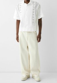 Bílá košile s krátkými rukávy a výřezy, kombinovaná s volnými, světle béžovými kalhotami a bílými teniskami; volný střih a prodyšný materiál.