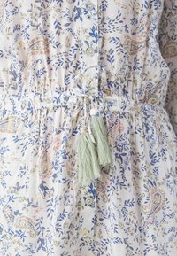 Hvit kjole med blå, rosa og beige blomstermønster i paisley, knappefront, innsnevret midje og to lysegrønne dusker knyttet rundt midjen.
