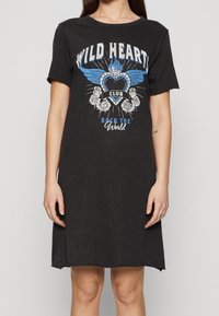 Nainen, jolla on päällä musta lyhythihainen mekko, jossa on sinivalkoinen sydän, siivet, ruusuja ja teksti "Wild Heart Club Rock The World."