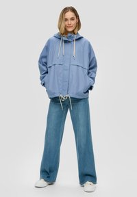 Veste légère bleu clair avec capuche, poches avant, taille ajustable à cordon de serrage et coupe décontractée, associée à un jean large.