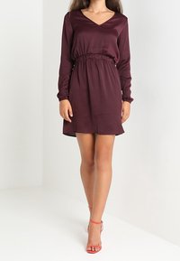 Mujer que lleva puesto un vestido de manga larga, de color púrpura oscuro, hasta la rodilla, con una cintura elástica y sandalias de tacón rojo, de pie frente a un fondo blanco.