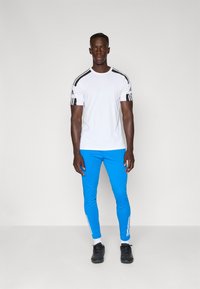 Hvid atletisk skjorte med sorte striber på skuldrene, parret med blå tætsiddende bukser med hvide sidestriber, iført sorte sko.