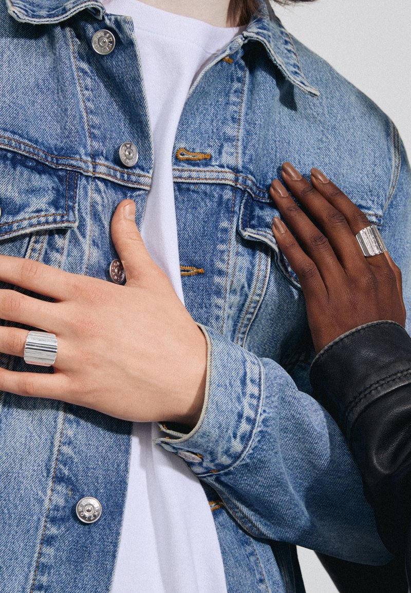 Giacca di denim con bottoni argentati, una maglietta bianca sotto, e due mani che indossano anelli argentati texture—uno con un motivo a righe.