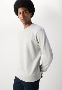 Light gray sweatshirt with a crew neck and ribbed cuffs, featuring a small square logo detail on the front. Soft fabric texture.