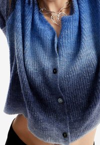 Blå stickad cardigan med en gradientdesign, strukturerad tyg och tre svarta knappar framtill. Mjuk och lätt.