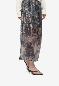 Jupe maxi en sequins argentés avec un motif dégradé, associée à des sandales noires à lanières. La jupe présente une silhouette fluide et des rangées de sequins verticaux.