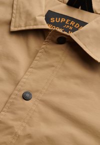 Tan overhemd met knoopsluiting en een spitse kraag, voorzien van een donkere knoop en een geweven label. De stof heeft een gladde textuur met een subtiele glans.