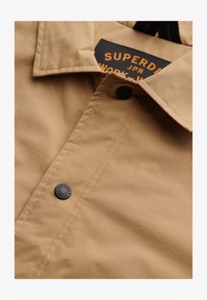 Beige Hemd mit spitzem Kragen, ausgestattet mit einem dunklen Knopf und einem gewebten Etikett. Der Stoff hat eine glatte Textur mit einem subtilen Glanz.