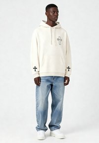 Felpa crema con grafica a croce nera sul petto e sui polsini delle maniche, abbinata a jeans azzurri chiari e sneaker bianche. Vestibilità rilassata e texture morbida.