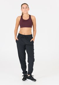 Bordeauxfarbener Sport-BH, schwarze Jogginghose mit elastischem Bund. Das Model steht selbstbewusst mit den Händen in den Taschen und trägt schwarze Sportschuhe.