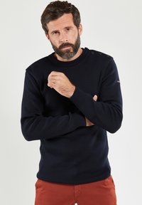 Marineblauer Strickpullover mit Rundhalsausschnitt und strukturiertem Stoff. Mit Schulterknöpfen und gerippten Bündchen ausgestattet. Kombiniert mit rötlichen Hosen.