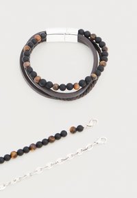 Bracelet en perles marron et noir avec des brins de cuir tressés et lisses, fermoir magnétique en argent, et deux petites chaînes argentées détachables avec des fermoirs.