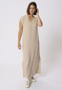 Beige Maxikleid mit V-Ausschnitt und kurzen Ärmeln, ausgestattet mit seitlichen Schlitzen. Kombiniert mit hellen Sneakers für einen Kontrast. Weicher Stoff.