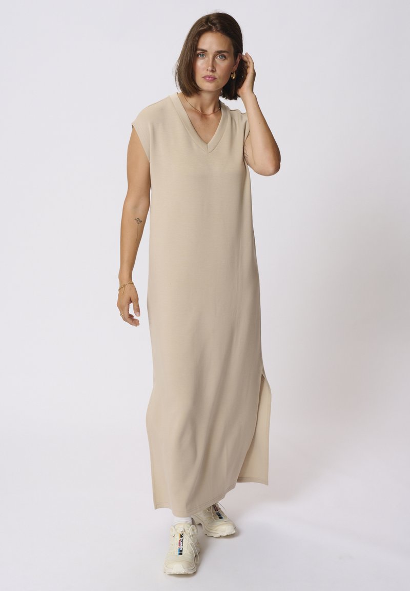 Beige Maxikleid mit V-Ausschnitt und kurzen Ärmeln, ausgestattet mit seitlichen Schlitzen. Kombiniert mit hellen Sneakers für einen Kontrast. Weicher Stoff.